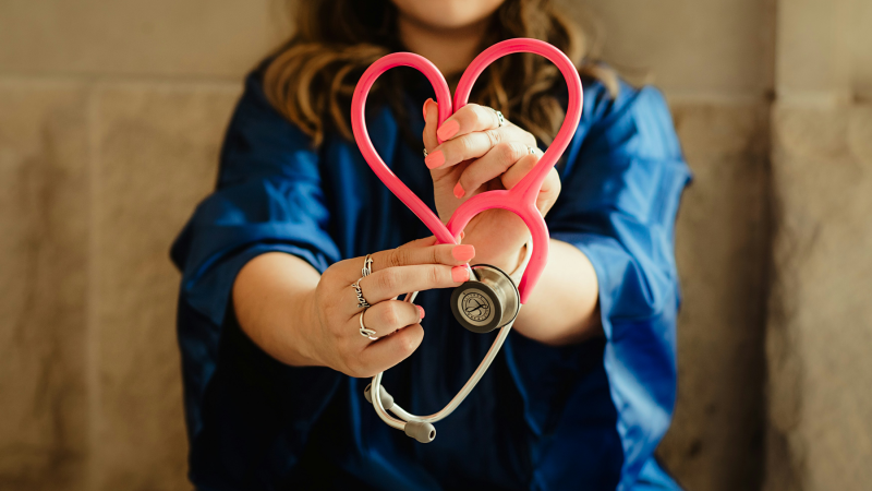 Close-up of female healthcare worker forming a heart shape with pink stethoscope.